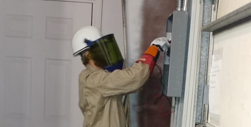 Person standing in a tan jumpsuit wearing  a facemask working on electrical work